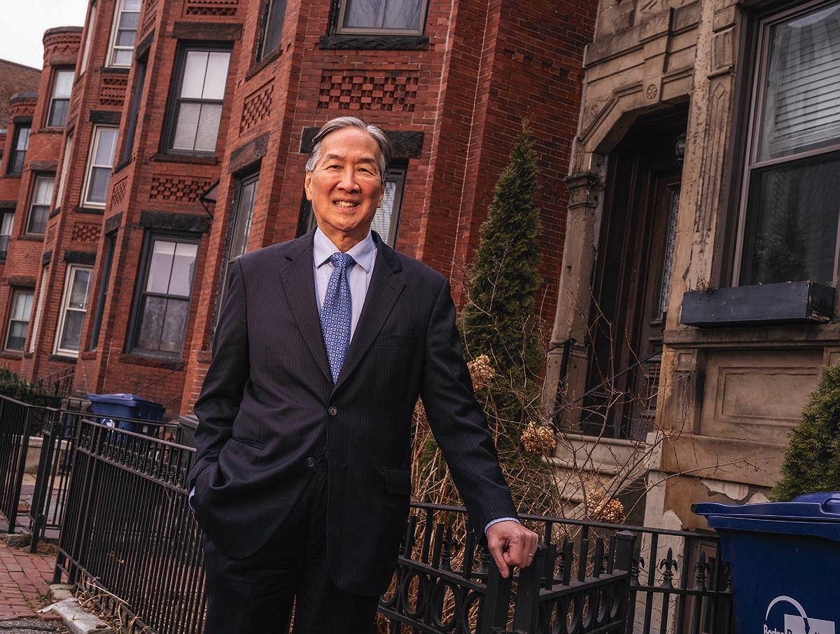 Howard Koh photographed on a sidewalk in Boston with homes behind him