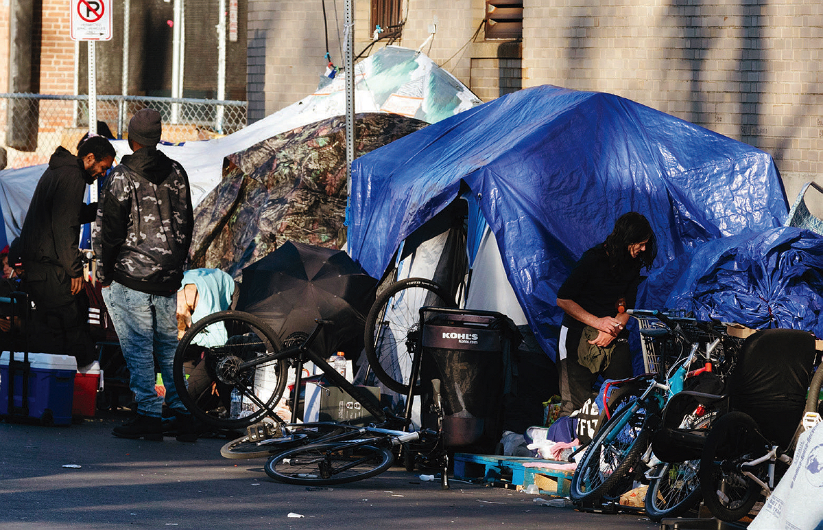 Three people stand beside several tents, tarps, and other possessions that line the sidewalk next to a brick building