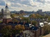 Skyline view of Harvard University with trees in autumn colors and a river under a cloudy sky.