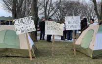 Tents and signs foreground a photograph of a rally in Allston organized by the Coalition for a Just Allston-Brighton on Tuesday, March 15