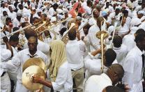 A street crowd of black men and women, all dressed in white, either playing or responding to the playing of dozens of trombones