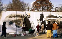 Along a city street, mural on a concrete wall shows two men shaking hands, with a large pair of watchful eyes in  the background.