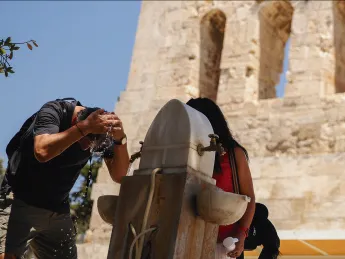 Man splashing water on his face at outdoor fountain beside woman holding cup near stone building.