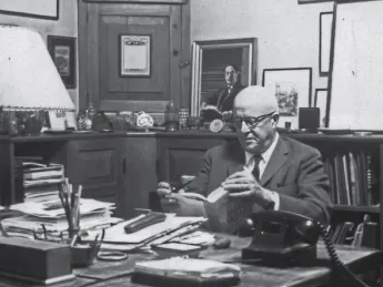Man in suit reading book at cluttered wooden desk in office filled with framed art and shelves.