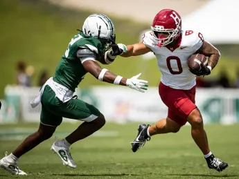 A football player in red dodges a tackle from a defender in green on a grassy field.