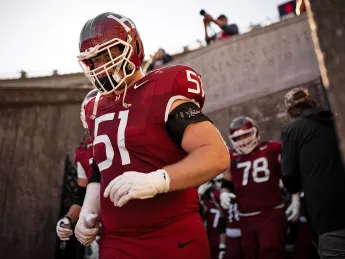 Harvard football player in red jersey 51 walks out of tunnel onto field with teammates behind.