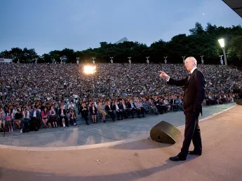 A speaker addresses a large crowd at an outdoor evening event.