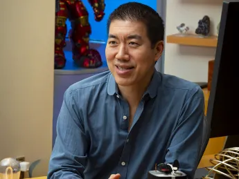 Professor David Liu smiles while sitting at a desk with colorful lanterns and a figurine in the background.