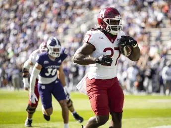 A football player in a red uniform runs with the ball, pursued by an opponent in a purple uniform.
