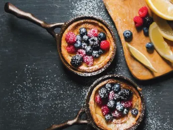 Two small cast iron pans with berry-topped desserts, dusted with powdered sugar, alongside lemon slices.