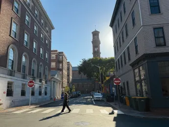A person walks across a street lined with historic buildings and a clock tower in the background.