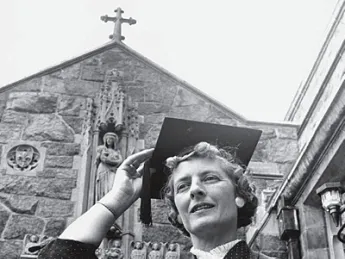 Woman in graduation cap smiling outdoors, church in the background, black and white photo.