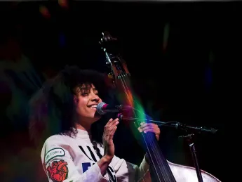 esperanza spalding, wearing a white jumpsuit with a "Life Force" logo, stands next to an upright bass