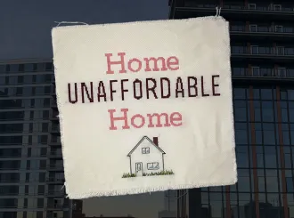 Cross-stitched fabric with the text "Home Unaffordable Home" over a background of high-rise apartment buildings.