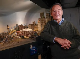 Frank Gevalt standing beside his detailed model city "Port Charles," featuring buildings, bridges, and an urban skyline.