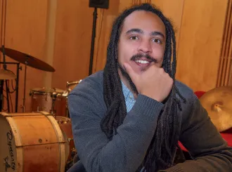 Jesse Cox smiling at a table with music notes and a book, with drums and cymbals behind him