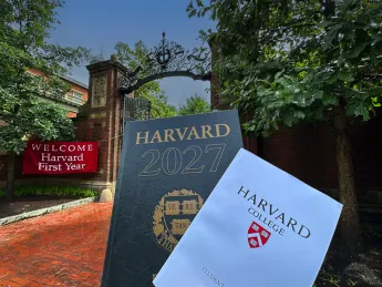 Harvard Gate in background with "Welcome Harvard First Year" red banner and in the foreground the Class of 2027 Class Book and Convocation program