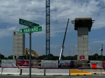 Construction site at the intersection of Academic Way and North Harvard Street. Two tall, unfinished concrete structures stand behind a fenced area with various signs and banners.