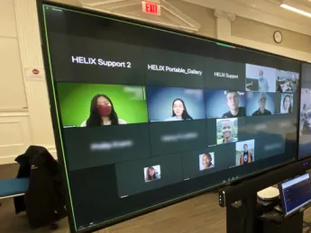 A hybrid classroom showing the large screens enabling a teacher to see all the students, remote or present, as he is followed by a camera operator.