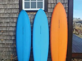 Two blue surfboards and an orange on rest on end against a weatherbeaten wall.