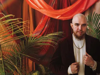 A formal portrait or José Olivarez in a gold chain and velvet suit jacket, standing amid red drapes and palm plants.