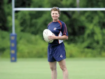 Holding a rugby ball, Sue Parker stands before rugby goalposts. She is Harvard&rsquo;s first varsity coach in the sport. 