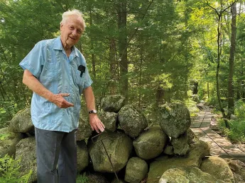 A white haired professor emeritus standing by a stone wall in the woods explaining its origins 