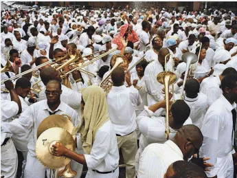 A street crowd of black men and women, all dressed in white, either playing or responding to the playing of dozens of trombones