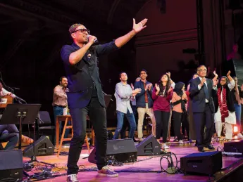 Singer Ali Sethi gestures toward the audience at Sanders Theatre, while students dance on stage behind him.