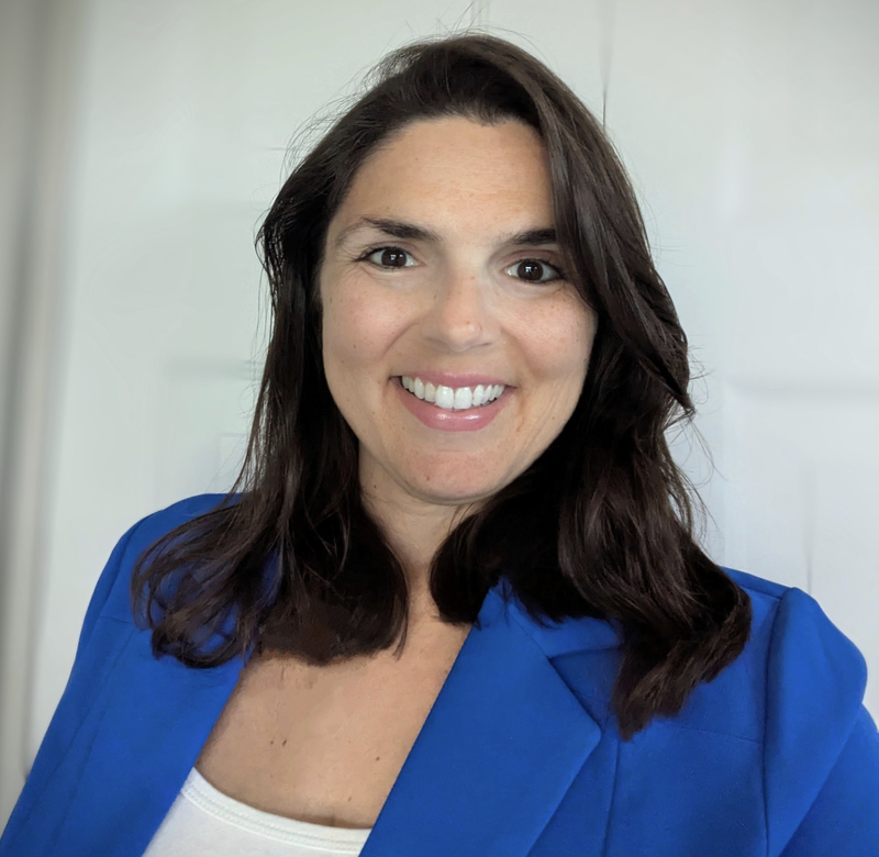 A woman with long dark brown hair and a bright smile is wearing a royal blue blazer over a white top. She is standing in front of a softly blurred white background, looking directly at the camera