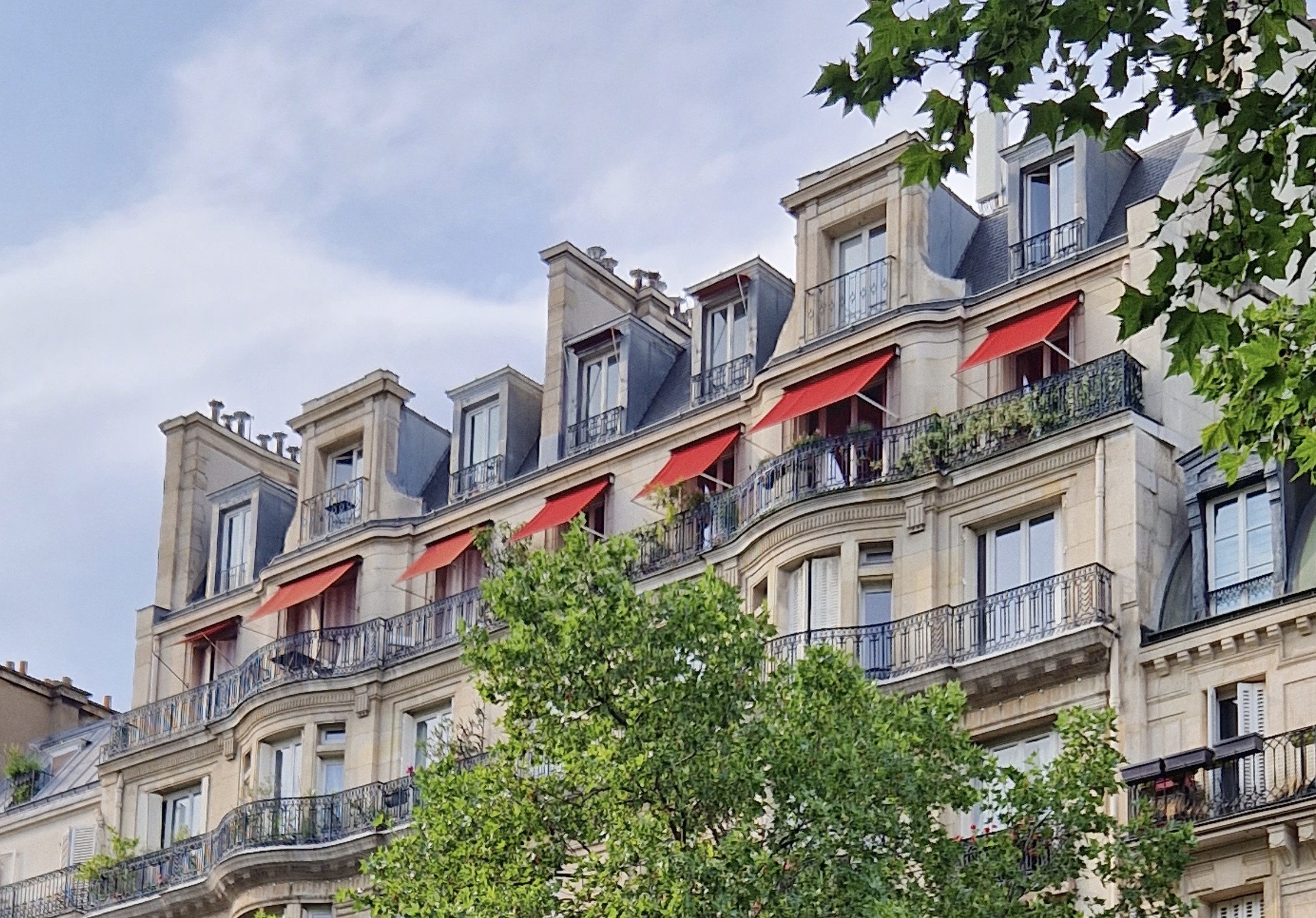  Elegant Parisian-style building with cream-colored stone, wrought-iron balconies, and red awnings, partially framed by green tree branches.