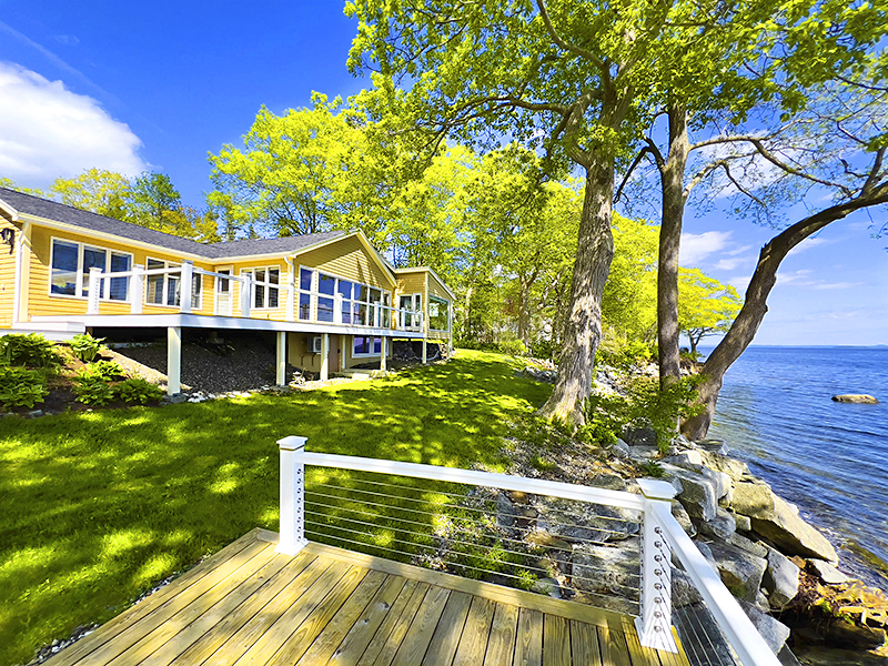 A bright yellow waterfront house with large glass windows sits elevated on stilts above a grassy lawn. Mature trees line the rocky shoreline, and a wooden deck with a white railing overlooks a calm, blue body of water under a sunny, clear sky.