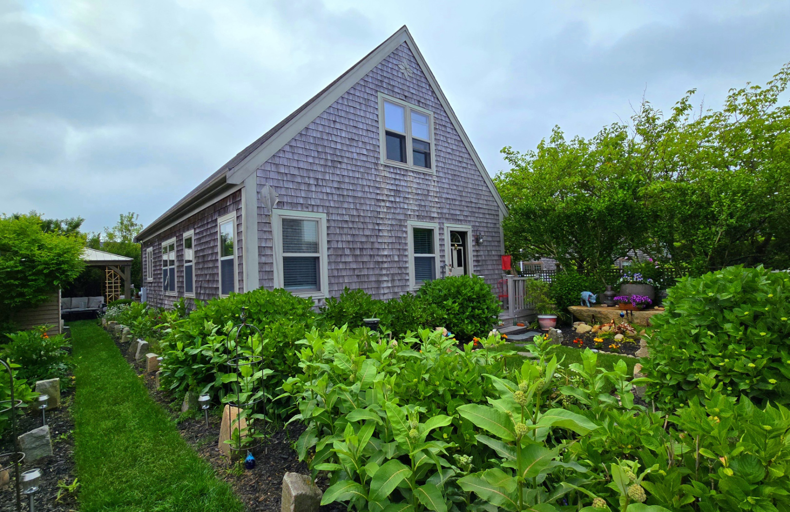 A charming cottage-style house with weathered gray shingles and a steep gabled roof, surrounded by a lush garden with leafy green plants, flowering shrubs, and a stone-edged pathway leading to a cozy gazebo.