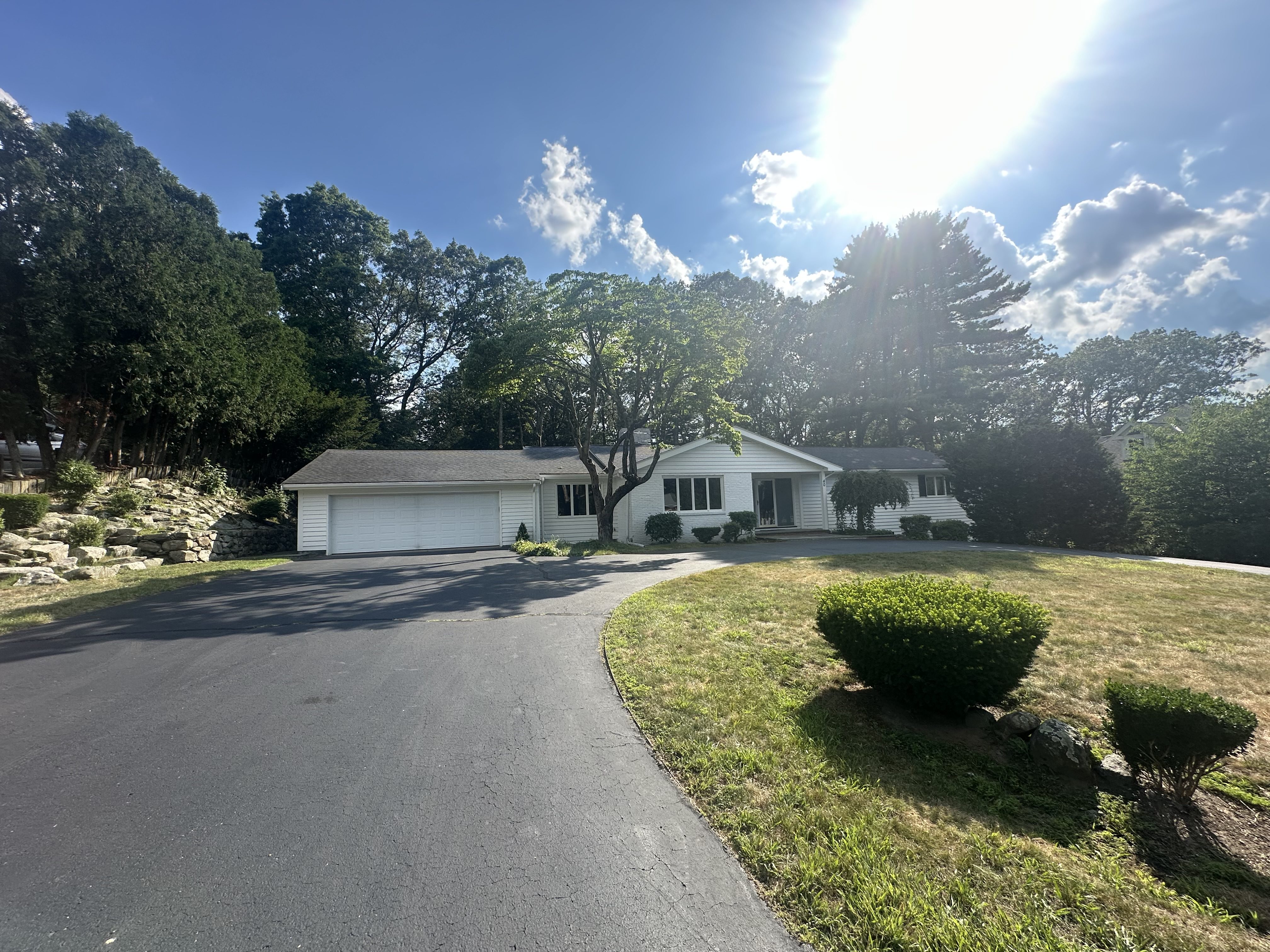 A single-story white house with a light gray roof and attached two-car garage sits in the center of the image. A smooth black asphalt driveway curves up to the house, flanked by a small round lawn with neatly trimmed shrubs. Tall trees and a rock wall line the left side of the property, while more trees form a wooded backdrop behind the house. The sun is shining brightly in a blue sky with scattered clouds, creating strong highlights and casting long shadows.