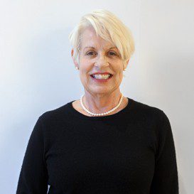 Smiling woman with short light hair, wearing a black top and pearl necklace, standing against a light background.