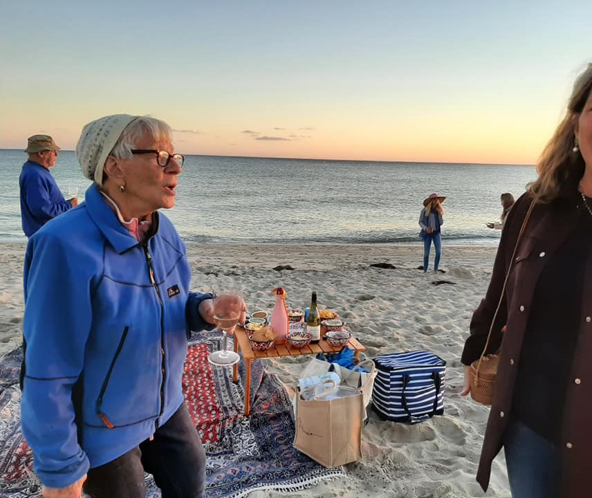 A joyful older woman in a blue jacket laughs on the beach at sunset, surrounded by friends.