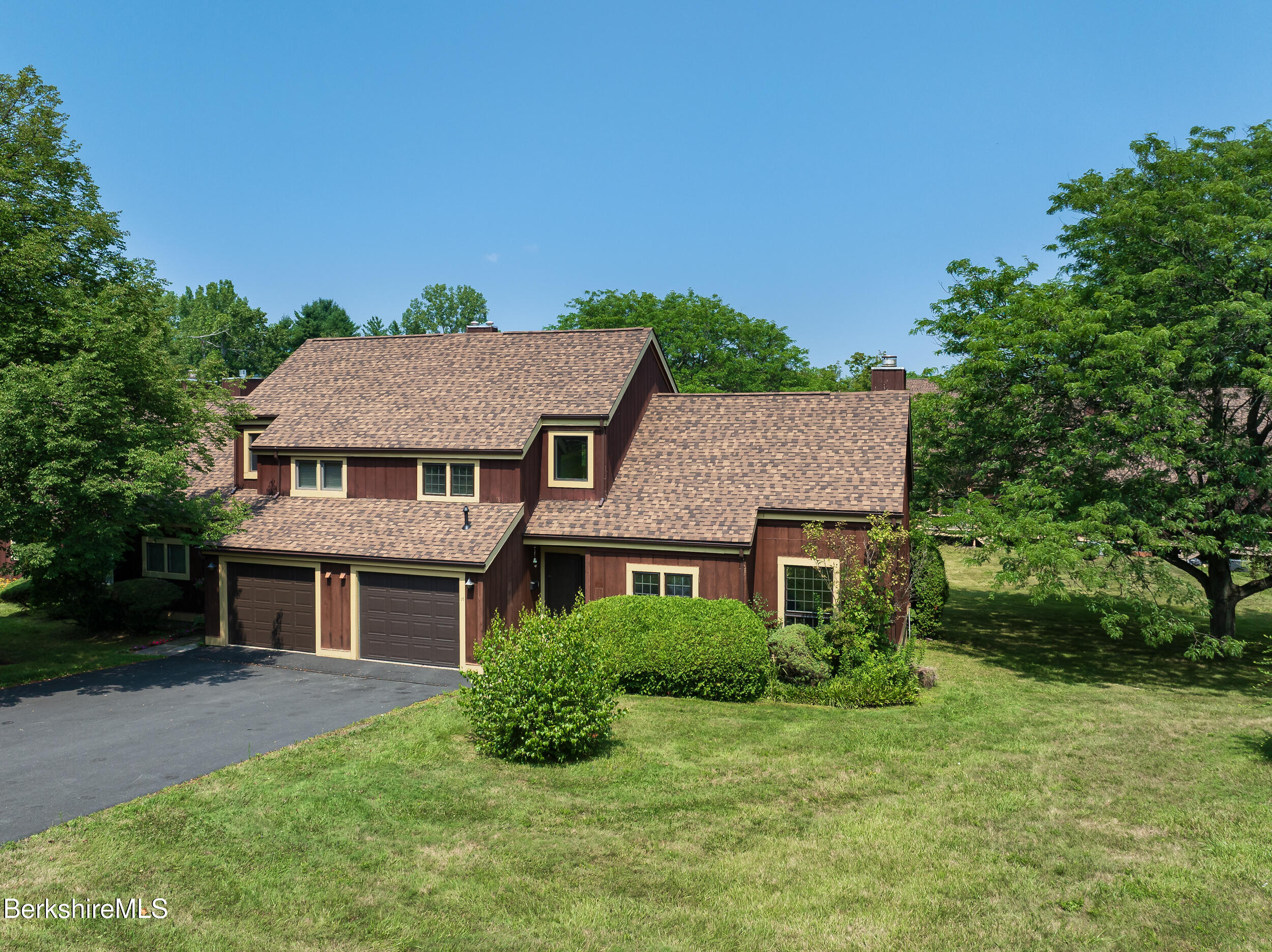 Two-story brown house with a driveway, surrounded by greenery and trees under a clear blue sky.