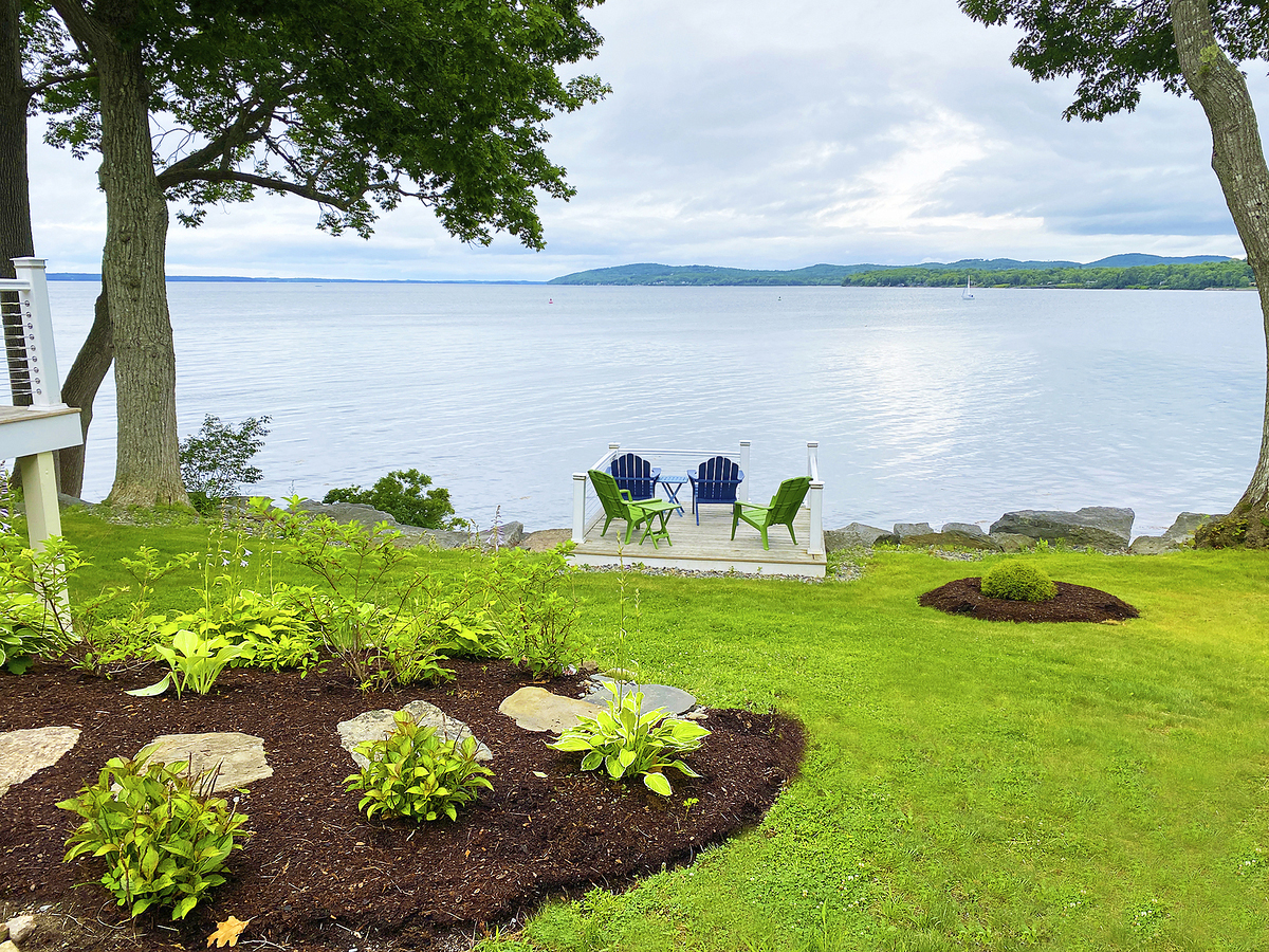 A green lawn with mulched flower/plant beds and trees overlooking the ocean. 