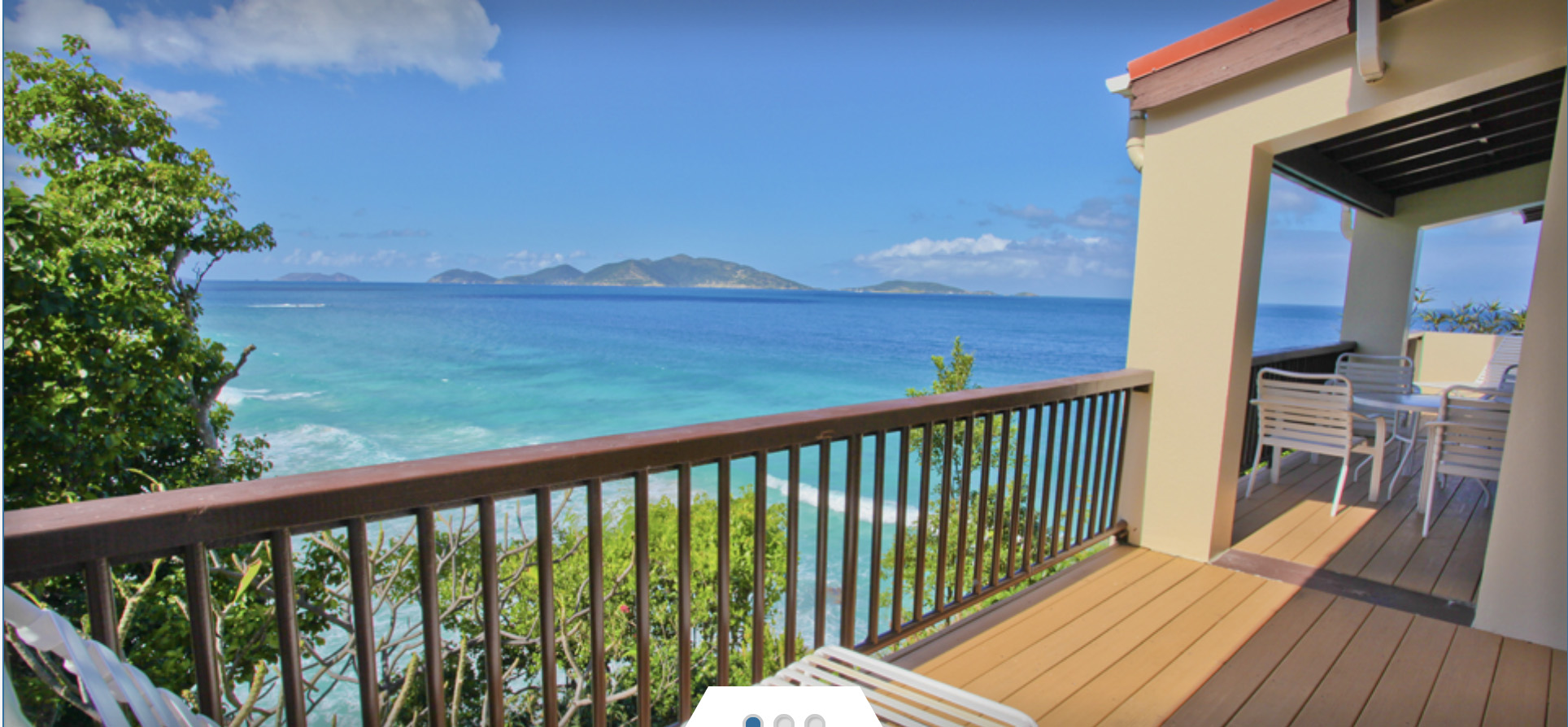 A wooden deck with a railing area and some white patio furniture overlooking the ocean.