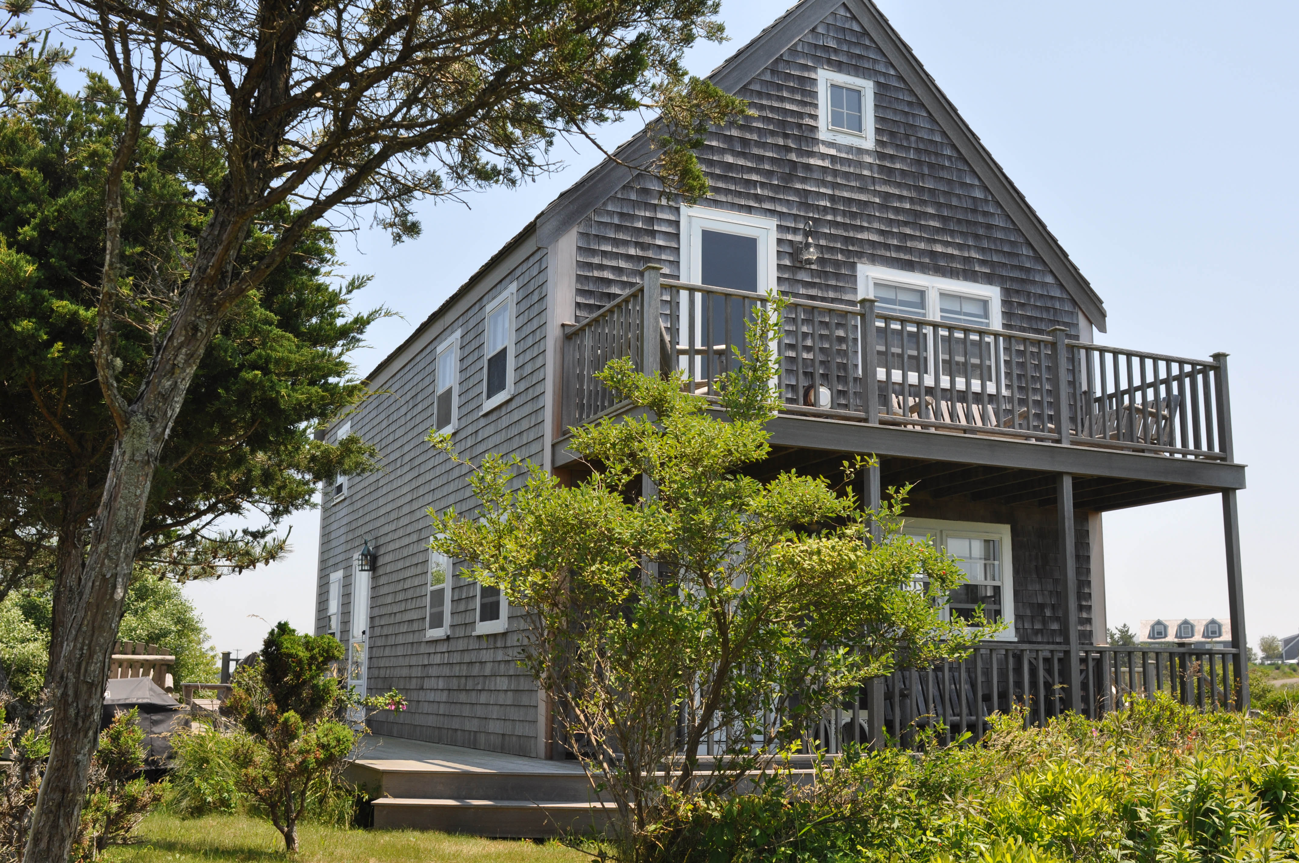 NANTUCKET DIONIS: Post and beam house, 5-minute walk to beach, adjacent to 350 acres of conservation land, 4 decks with extensive views, 3 bedrooms, 2 bathrooms, high-speed internet. Book early for ferry reservations. View photos: http://bit.ly/dionisma. 530-574-7724, jdhamilton@outlook.com or 530-574-7731, doctorpaula@comcast.net. Photo of a two-story gray house with trees in the background.