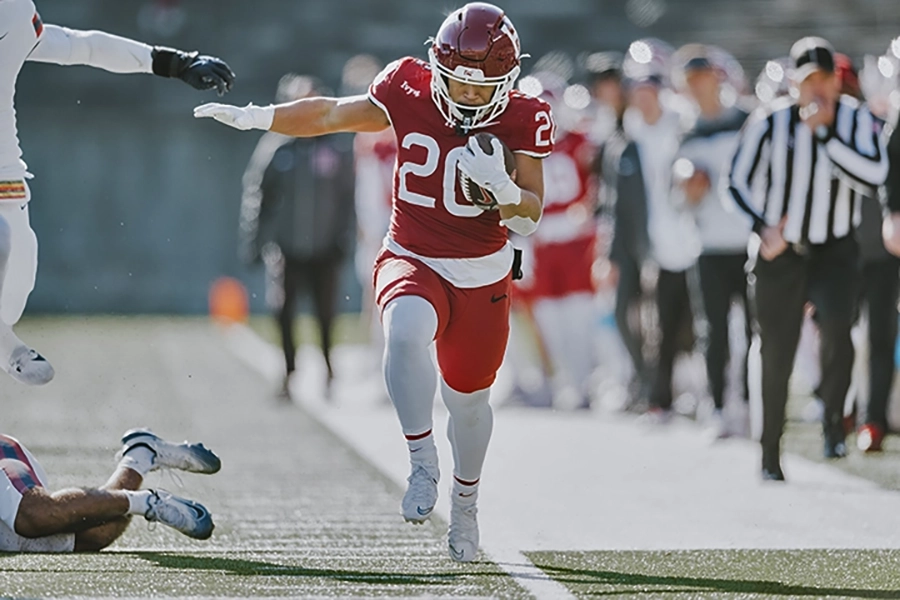 Harvard player in red jersey runs down the sideline with the football, evading a diving defender.