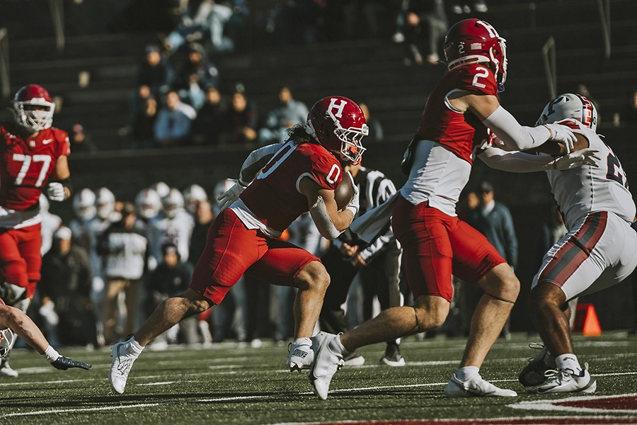 Harvard running back in red jersey carries the ball while teammates block opposing defenders.