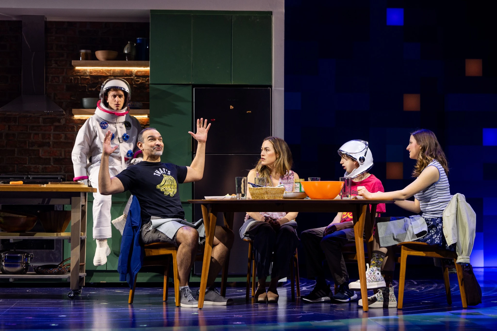 Members of the cast of Wonder sit at a table together having a conversation in a scene.