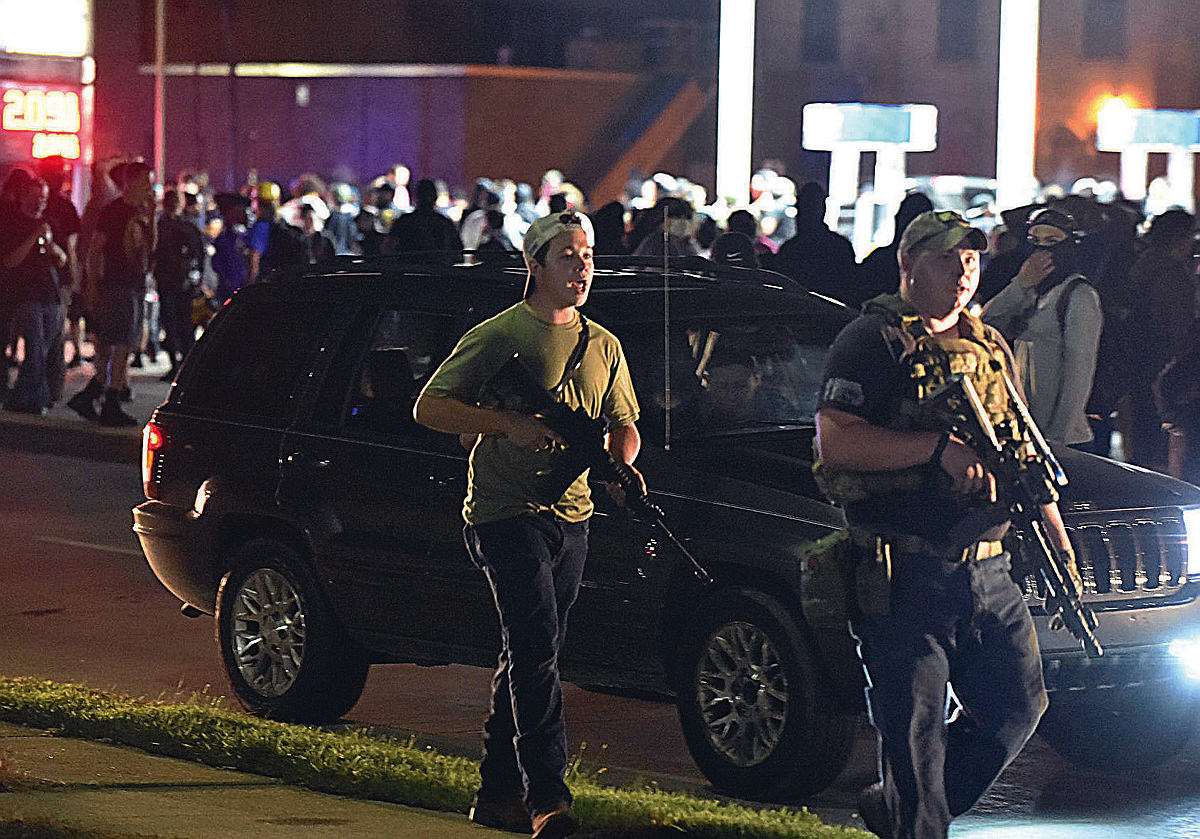 Wearing a backwards cap and carrying an AR-15-style rifle, Kyle Rittenhouse walks down a street at night in front of protesters. 