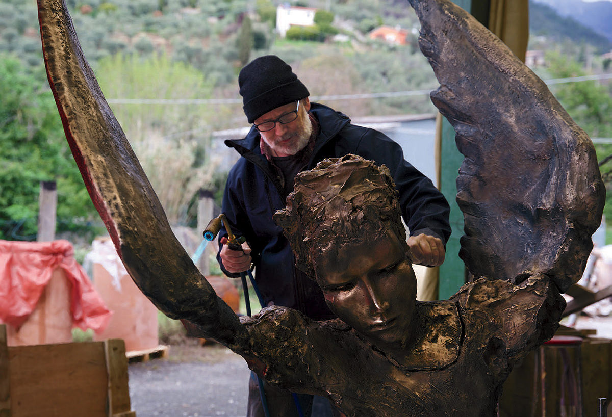 Del Deo stands behind a large sculpted face and winged torso, working with a blowtorch and a blade.