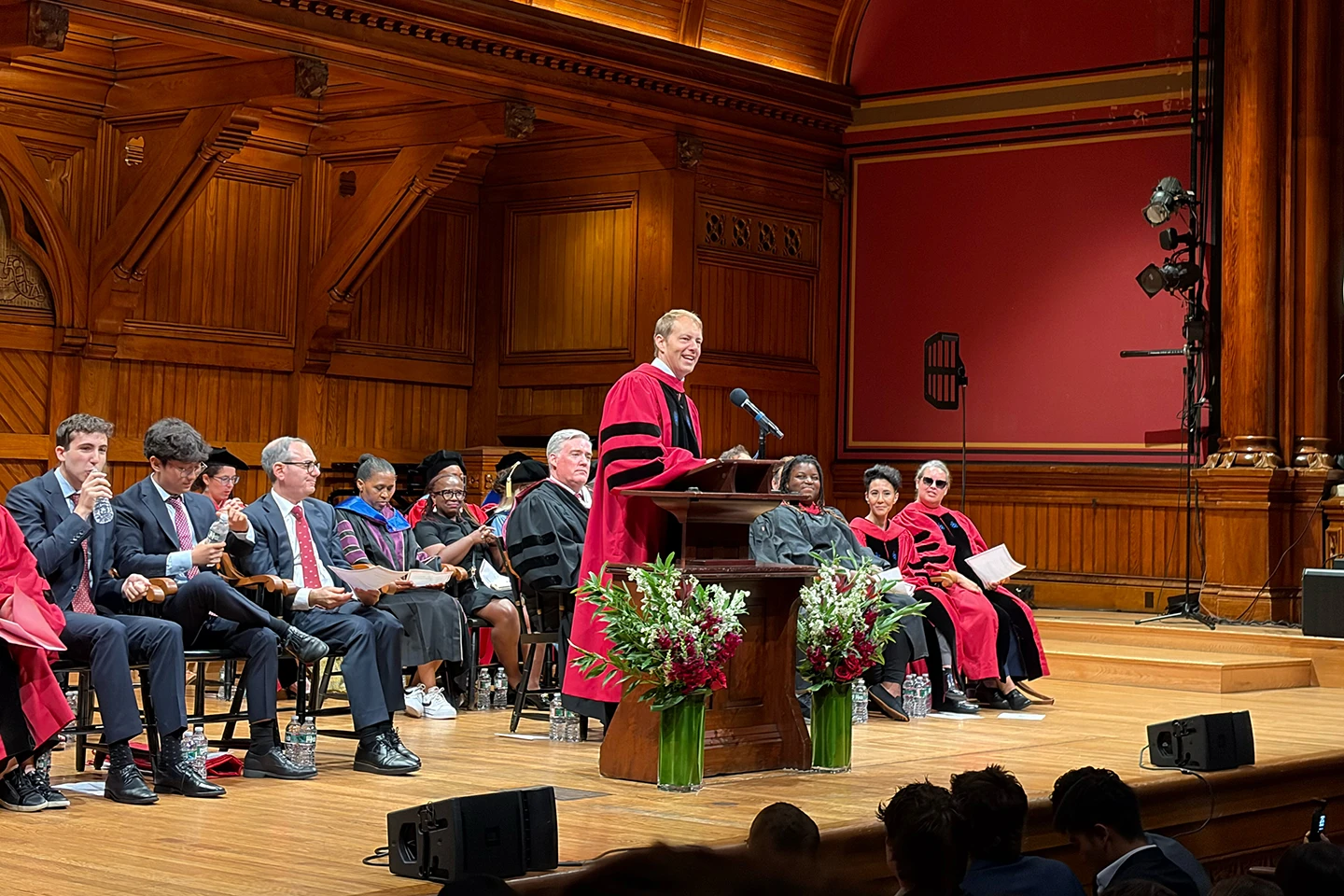 A man wearing a red and black robe speaks into a microphone at a podium while a crowd sits behind him on a stage