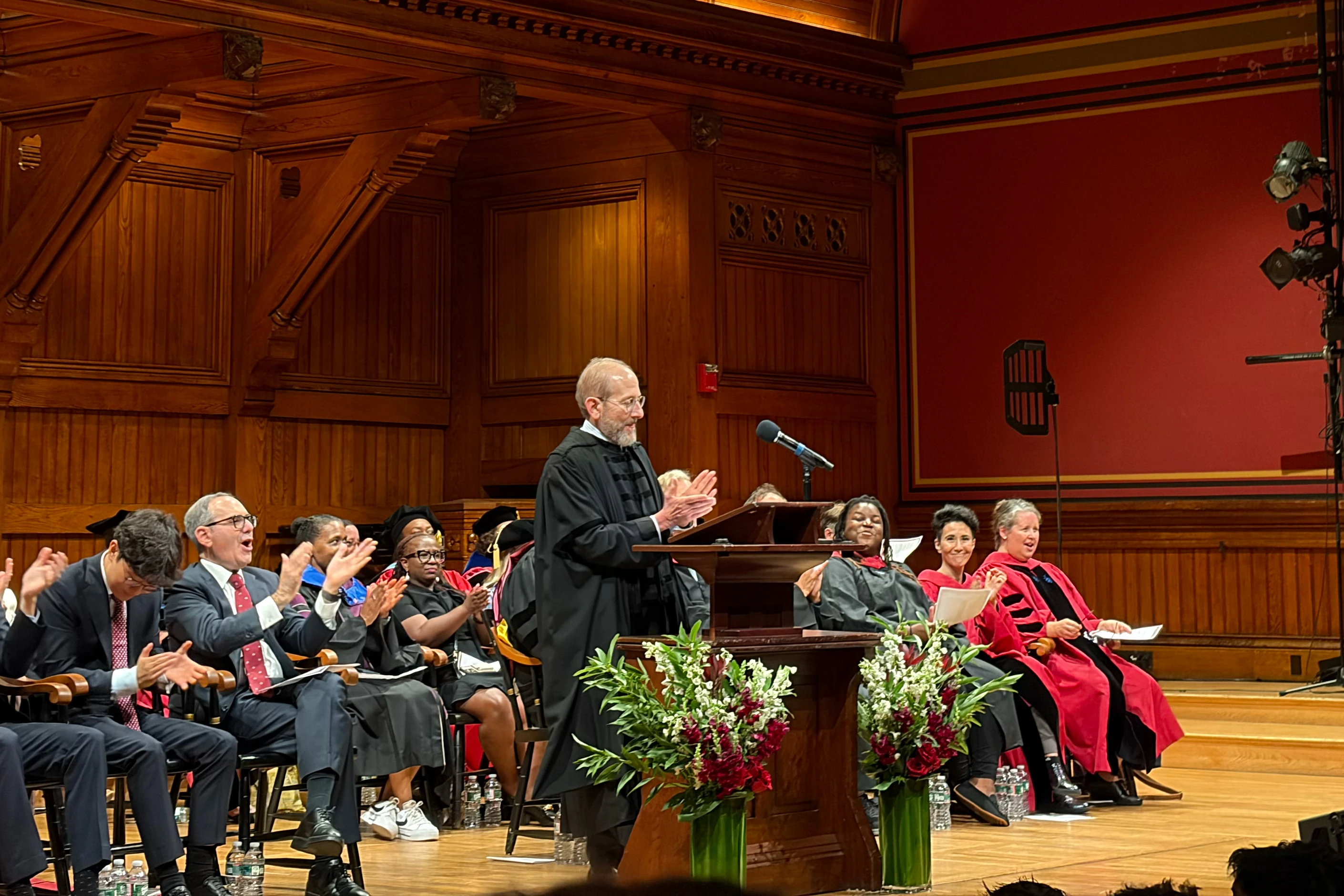 A man in a black robe claps while speaking at a podium inside a wood paneled room in front of a crowd of people sitting on a stage