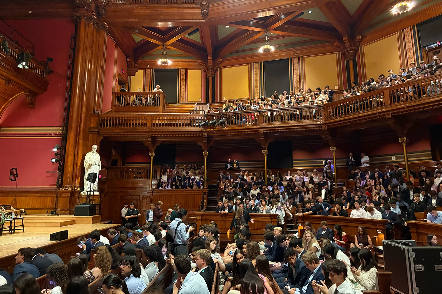 A crowd of people sit in an auditorium with wooden risers and balconies