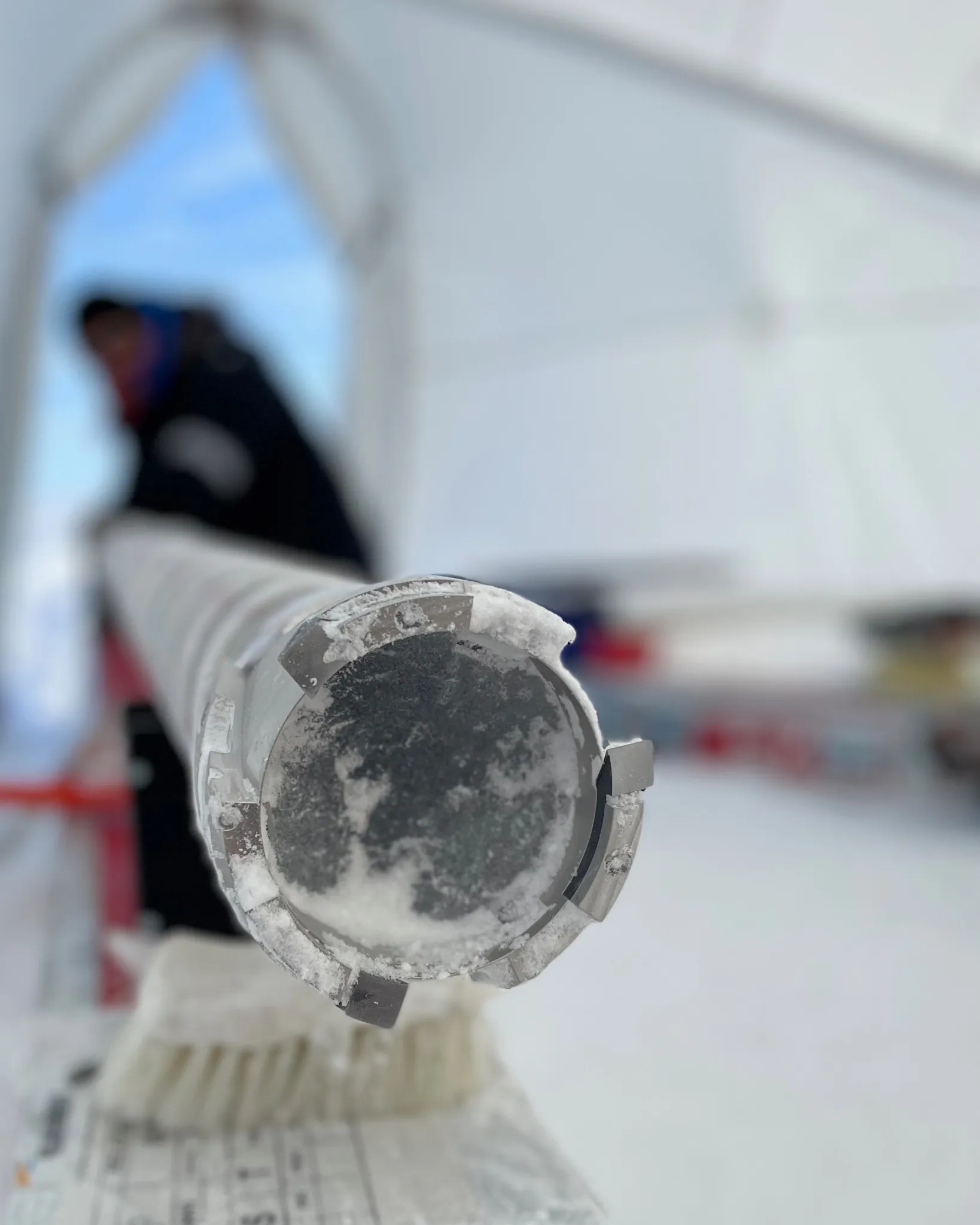 A frosted ice-core drill head in focus with a researcher blurred behind it.