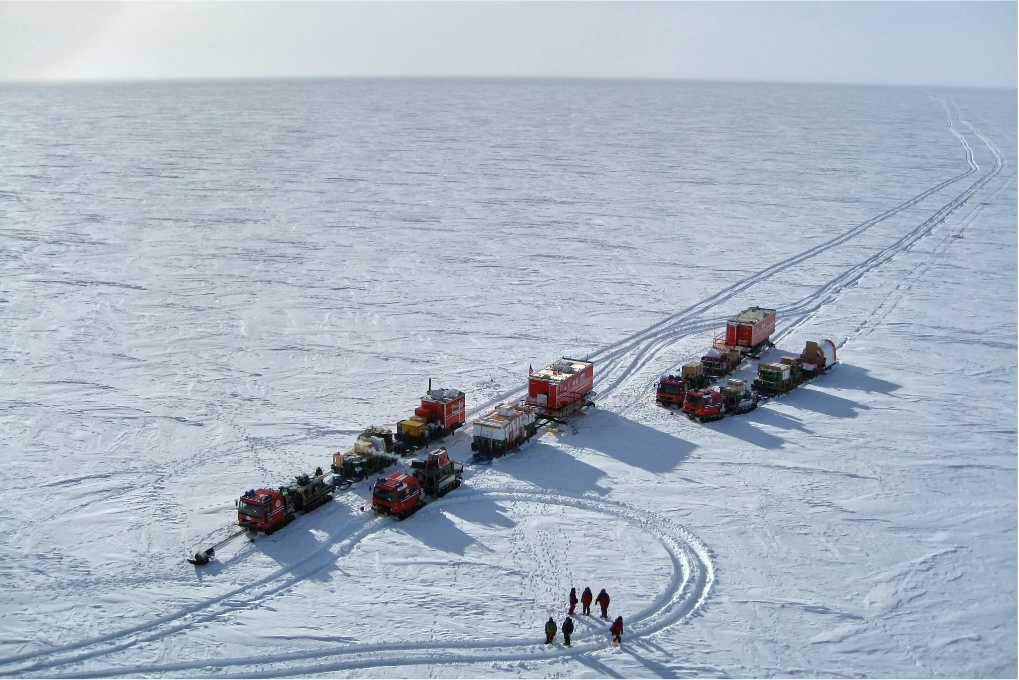 An aerial view of an expedition vehicle convoy crossing a vast snowy ice sheet.